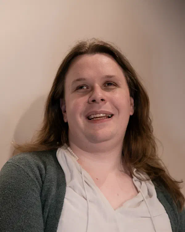 Shane Dittmar, a white, hazel-eyed person with long blonde hair, smiles at the camera with one arm crossed over their body. They wear a white shirt under a dark green cardigan.